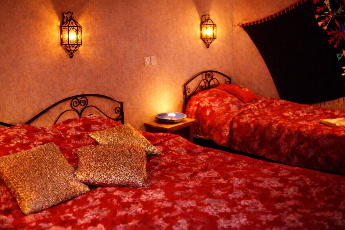 Habitación de hotel con dos camas, cada una con ropa de cama y almohadas decorativas de motivos florales rojos. Hay apliques de pared y una mesita de noche con lámpara.