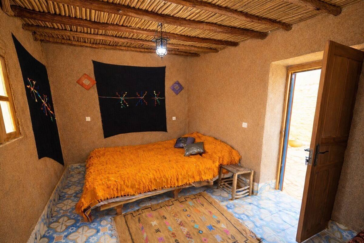 Una habitación con una cama, un taburete de madera, una puerta y un tapiz en la pared.