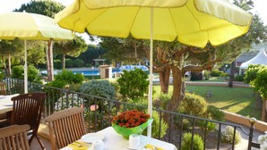 An outdoor dining area with yellow umbrellas, a table set with plates, a bowl of red flowers, and a view of a pool and greenery. - Hotel La Romarine (Ramatuelle)