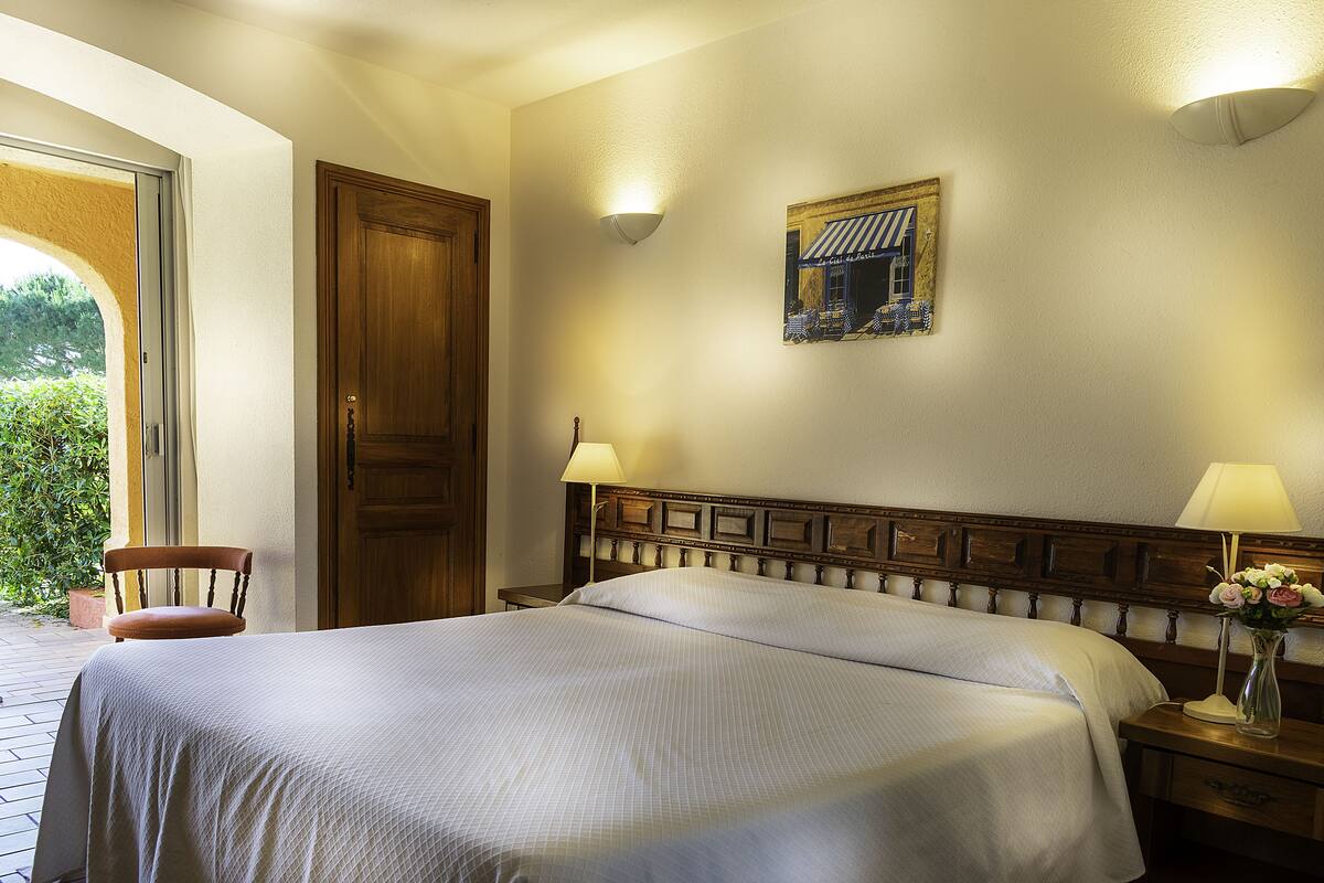 Una cama bien hecha con tablero de madera, una silla de madera y dos lámparas de noche.