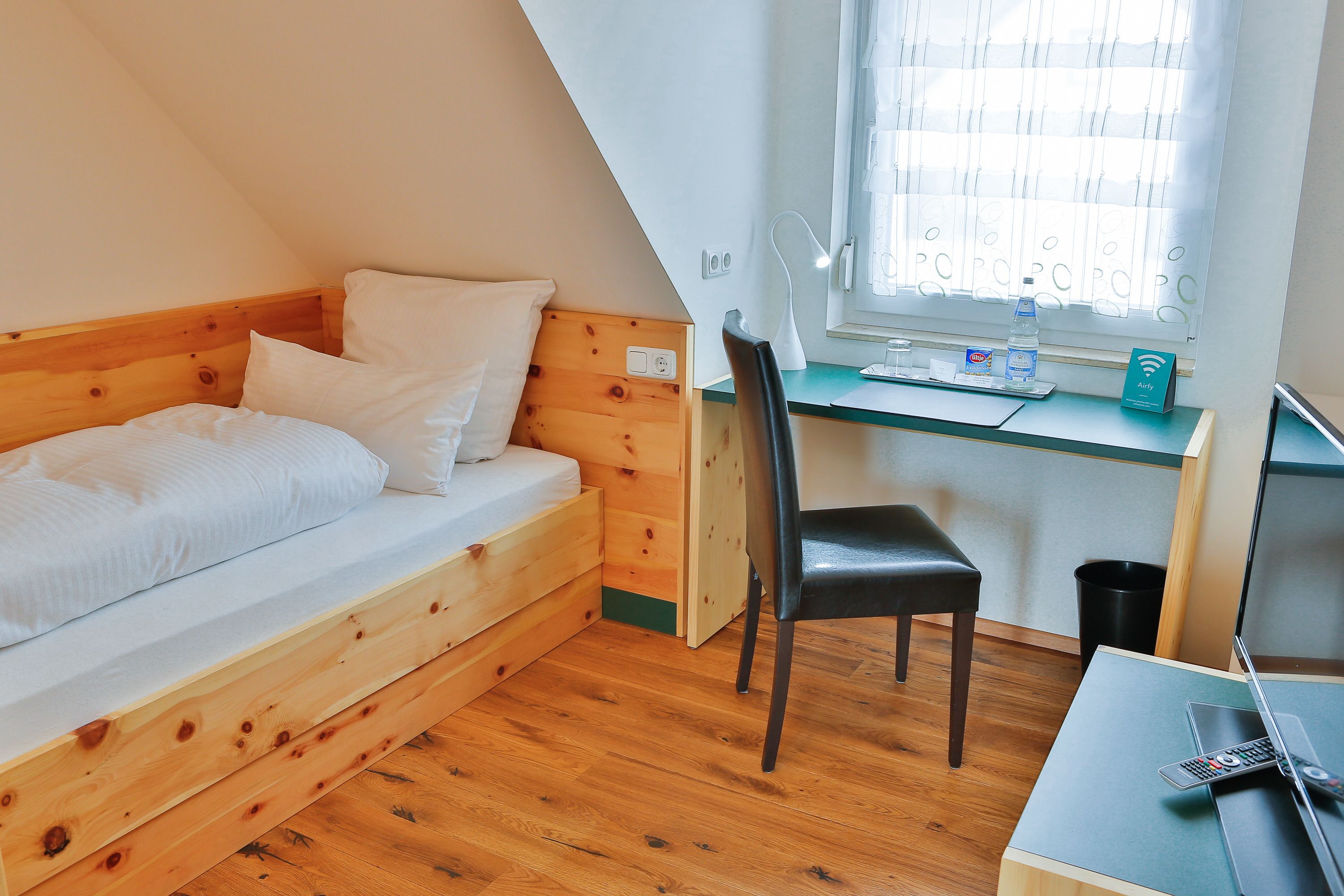 A small, cozy room with a wooden bed, a desk with a laptop, a chair, and a window with blinds.