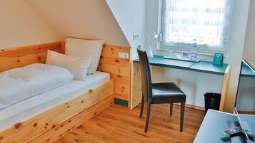 A small, cozy room with a wooden bed, a desk with a laptop, a chair, and a window with blinds.