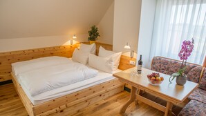 Una cama de madera con ropa de cama blanca, una mesita con fruta y un florero, y un sofá estampado con motivos florales.