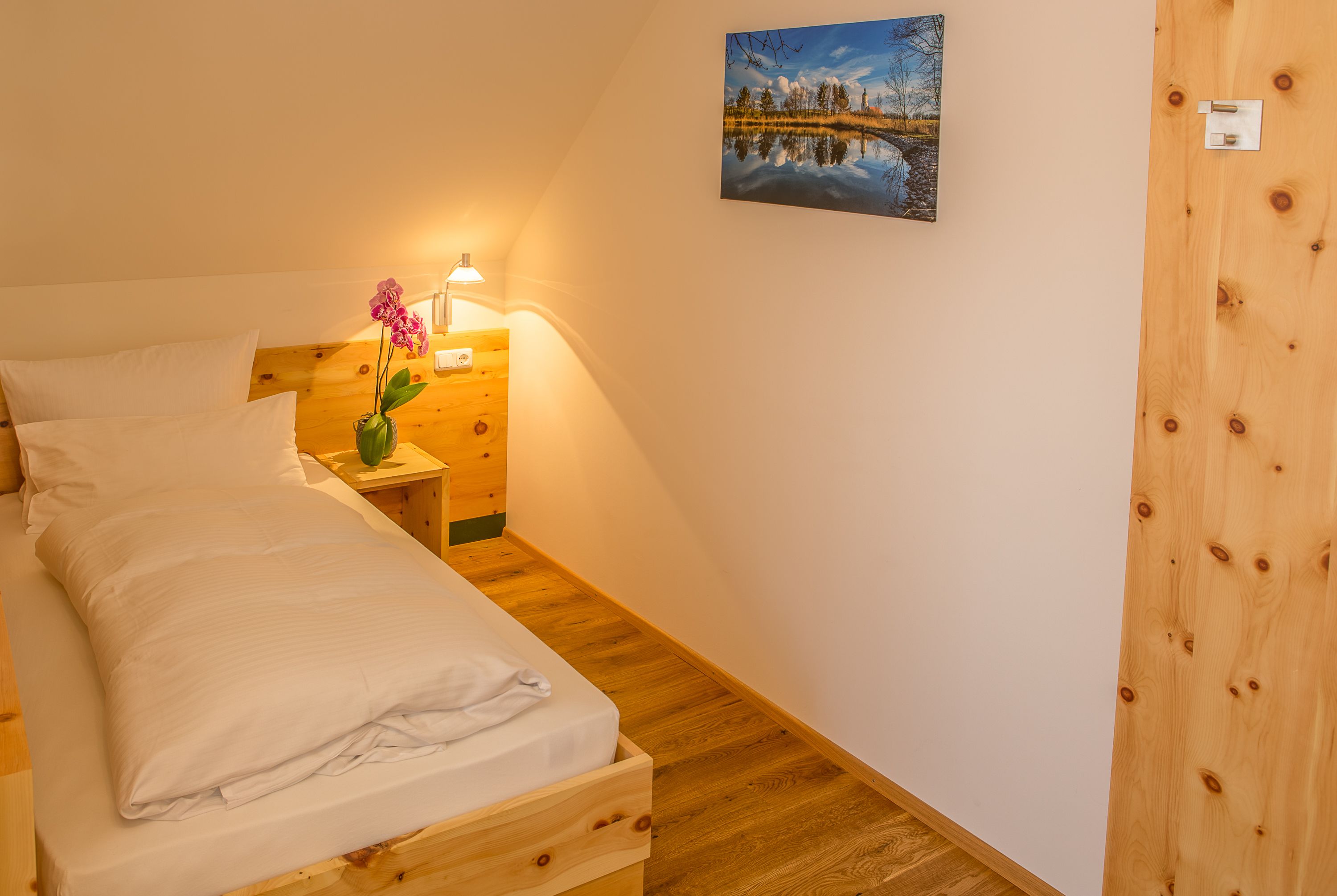 Een net opgemaakt bed met een houten hoofdeinde, een nachtkastje met een lamp, een vaas met bloemen en een schilderij aan de muur.