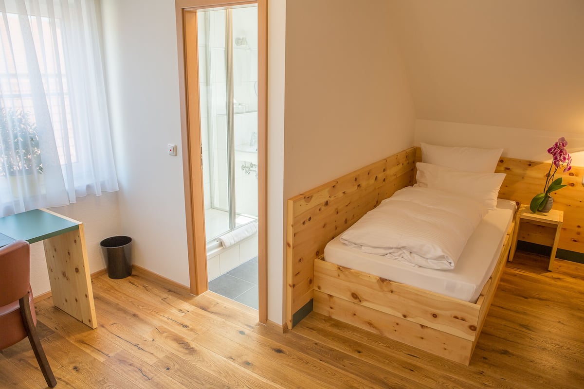 Una cama de madera con ropa de cama blanca, una mesita de noche con una planta y una silla en una habitación con suelo de madera y una ventana con cortinas de encaje.