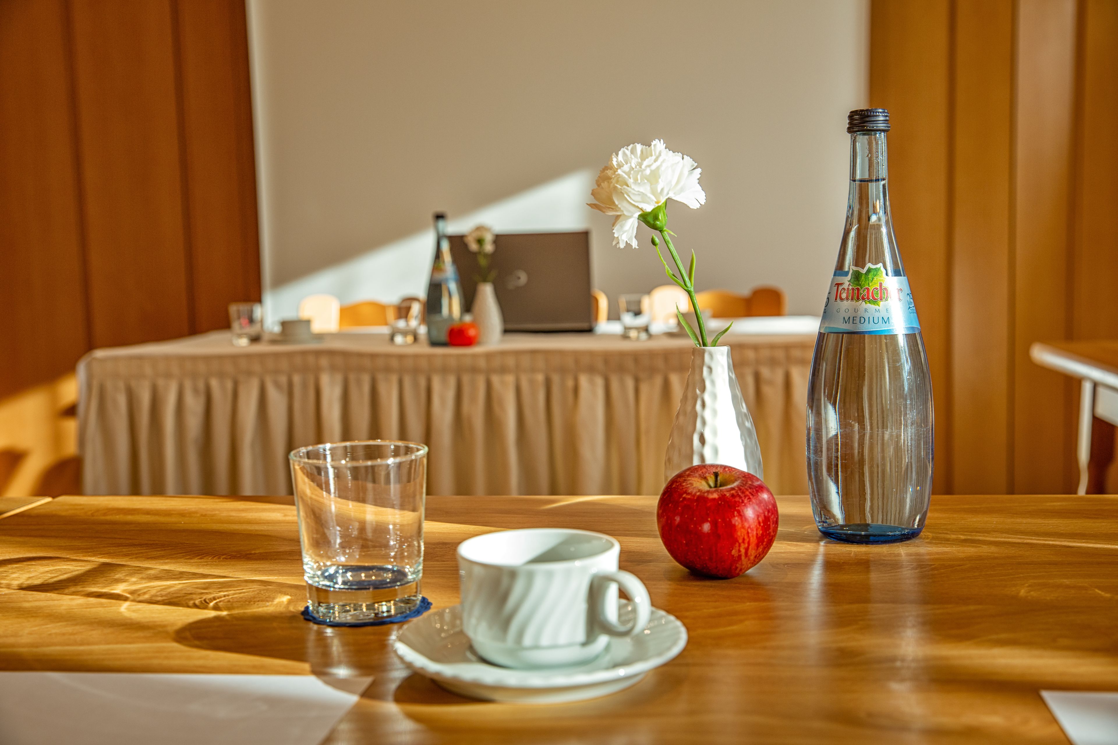 Une table en bois sur laquelle se trouvent un verre d’eau, une tasse blanche, une pomme rouge, une fleur blanche dans un vase et une bouteille de vin rouge Teimacher.