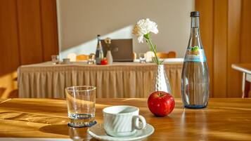 Une table en bois sur laquelle se trouvent un verre d’eau, une tasse blanche, une pomme rouge, une fleur blanche dans un vase et une bouteille de vin rouge Teimacher.