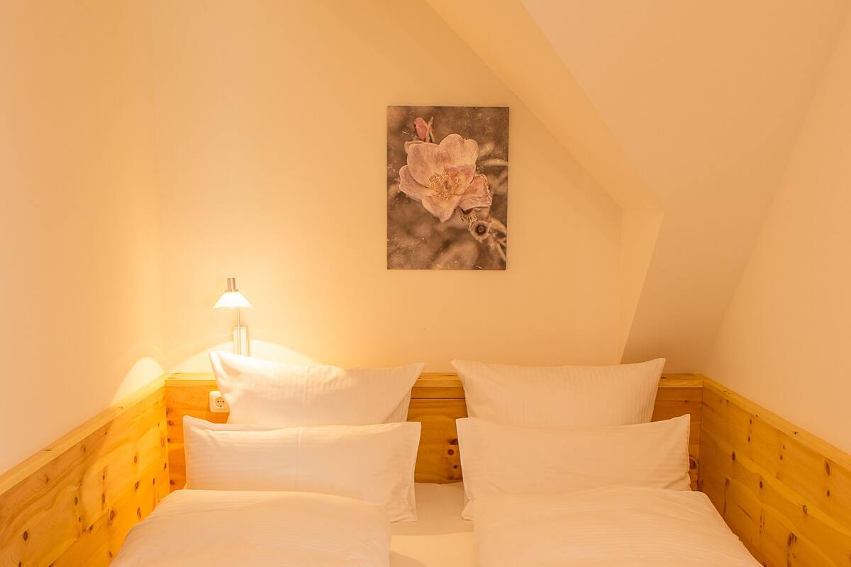 Una cama individual con cabecera y piecera de madera, ropa de cama blanca, una mesita de noche con lámpara, y un cuadro en la pared.