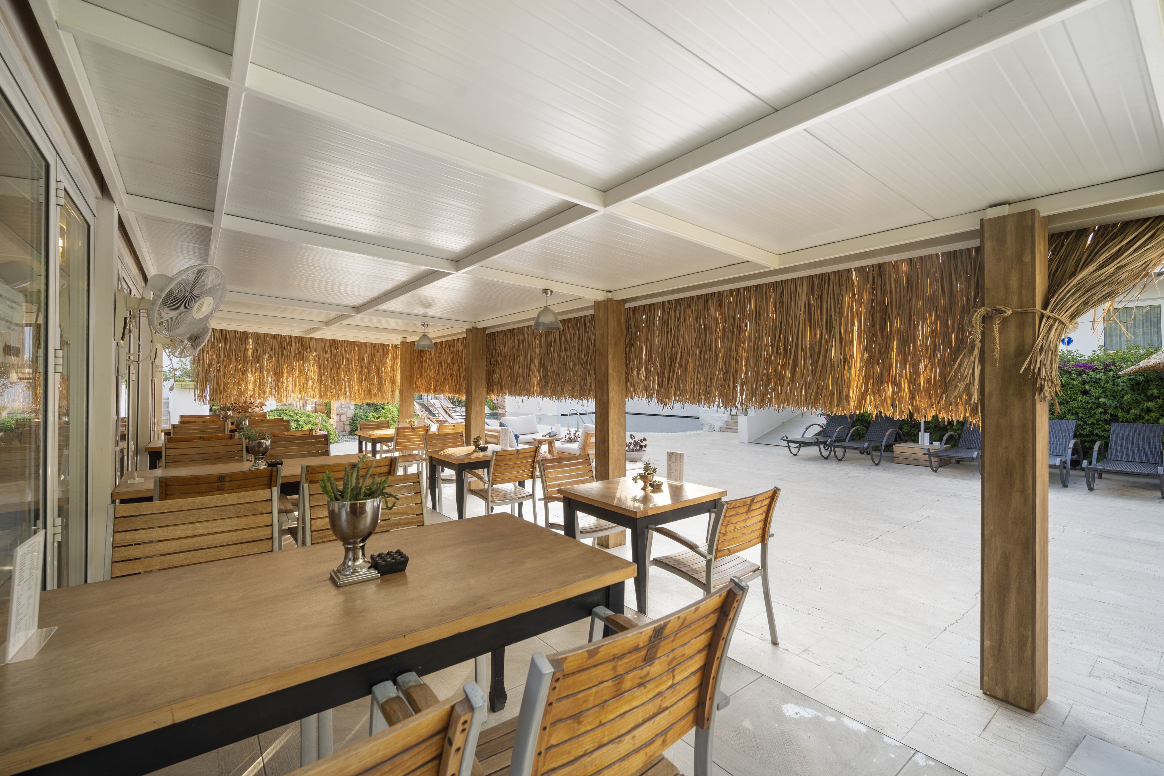 Un restaurante al aire libre con mesas y sillas de madera, un ventilador y un techo de paja.