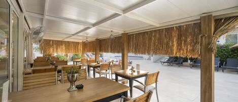 Un restaurante al aire libre con mesas y sillas de madera, un ventilador y un techo de paja.