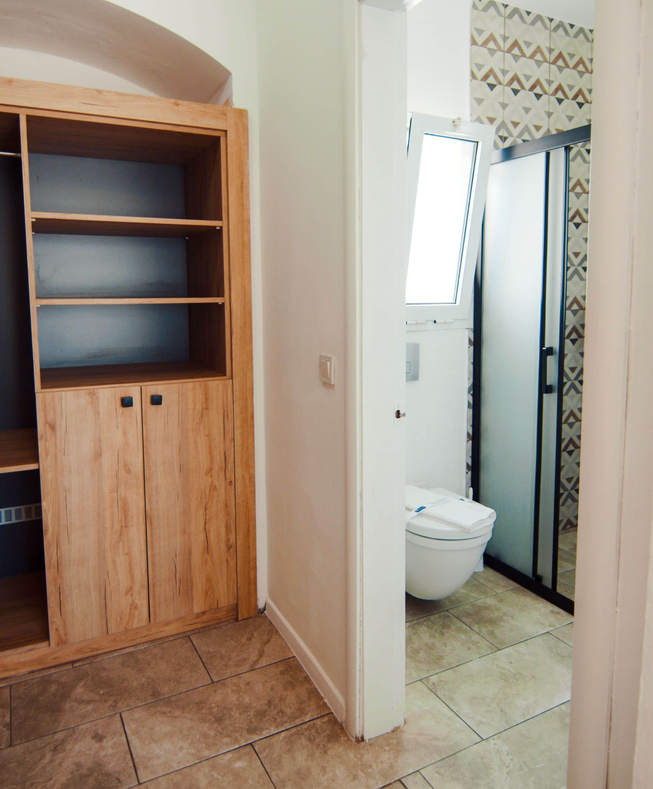 A bathroom with a toilet, a shower, and a wooden wardrobe with multiple shelves.