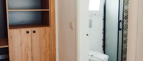 A bathroom with a toilet, a shower, and a wooden wardrobe with multiple shelves.