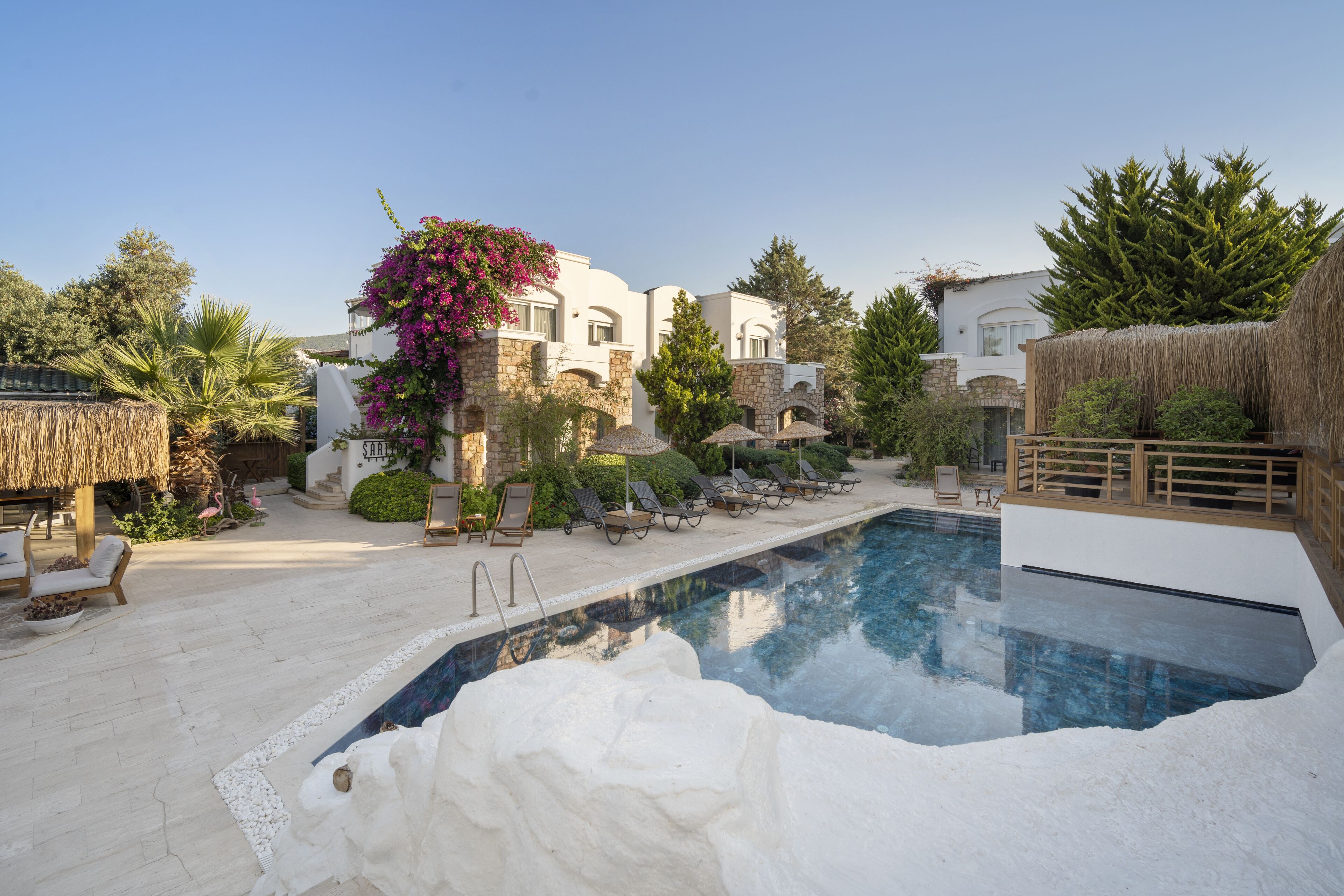 Un espace piscine avec des chaises longues, un parasol en chaume et un bâtiment en pierre doté d’une façade blanche.