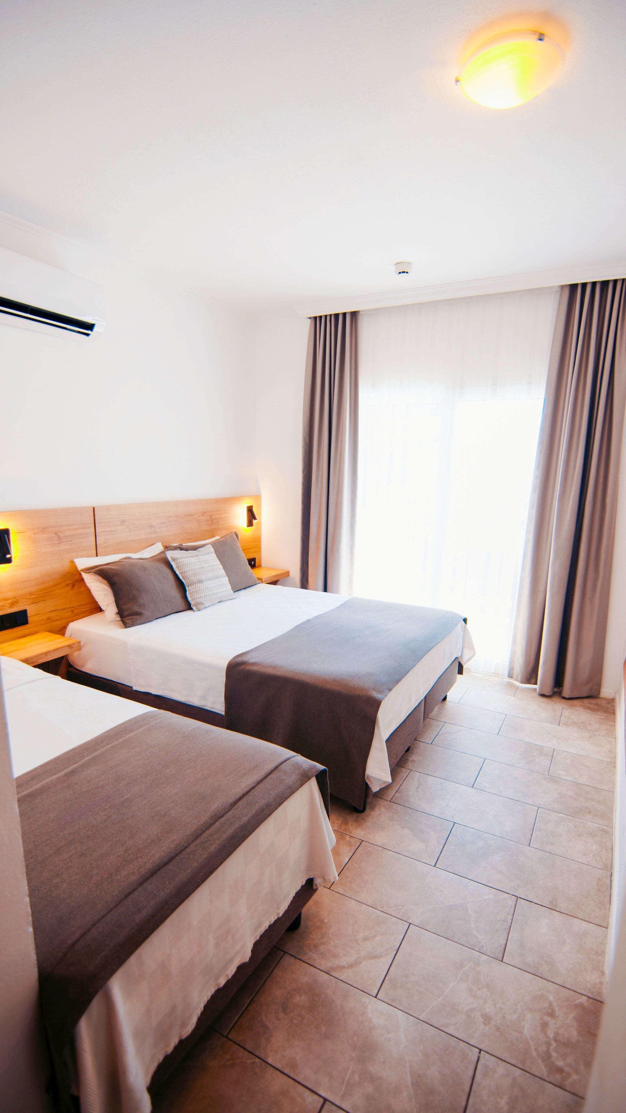 A hotel room with two beds, a wooden headboard, a nightstand, a wall-mounted light, and a window with curtains.