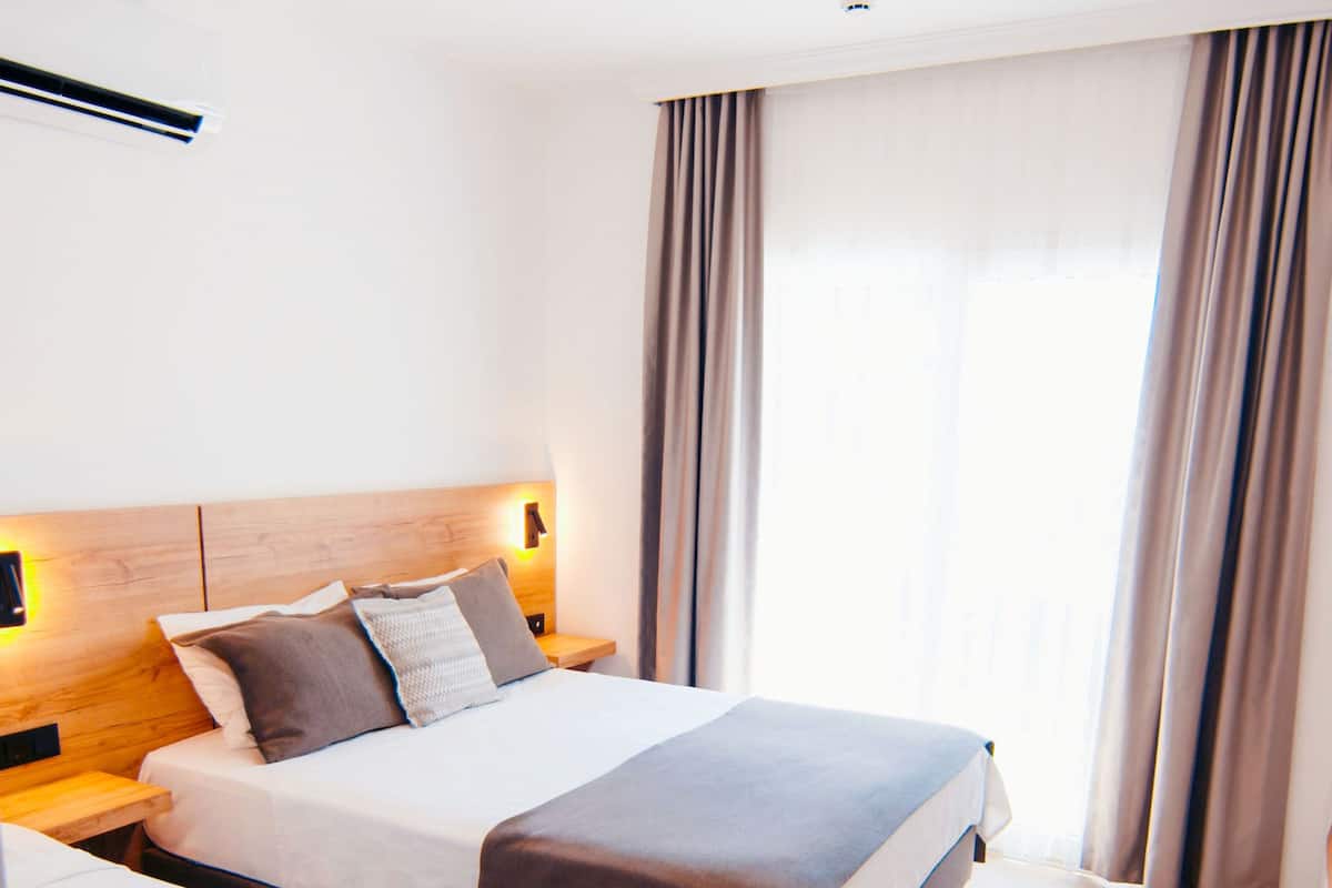 Habitación de hotel con dos camas, cabecera de madera, mesita de noche, lámpara de pared y ventana con cortinas.