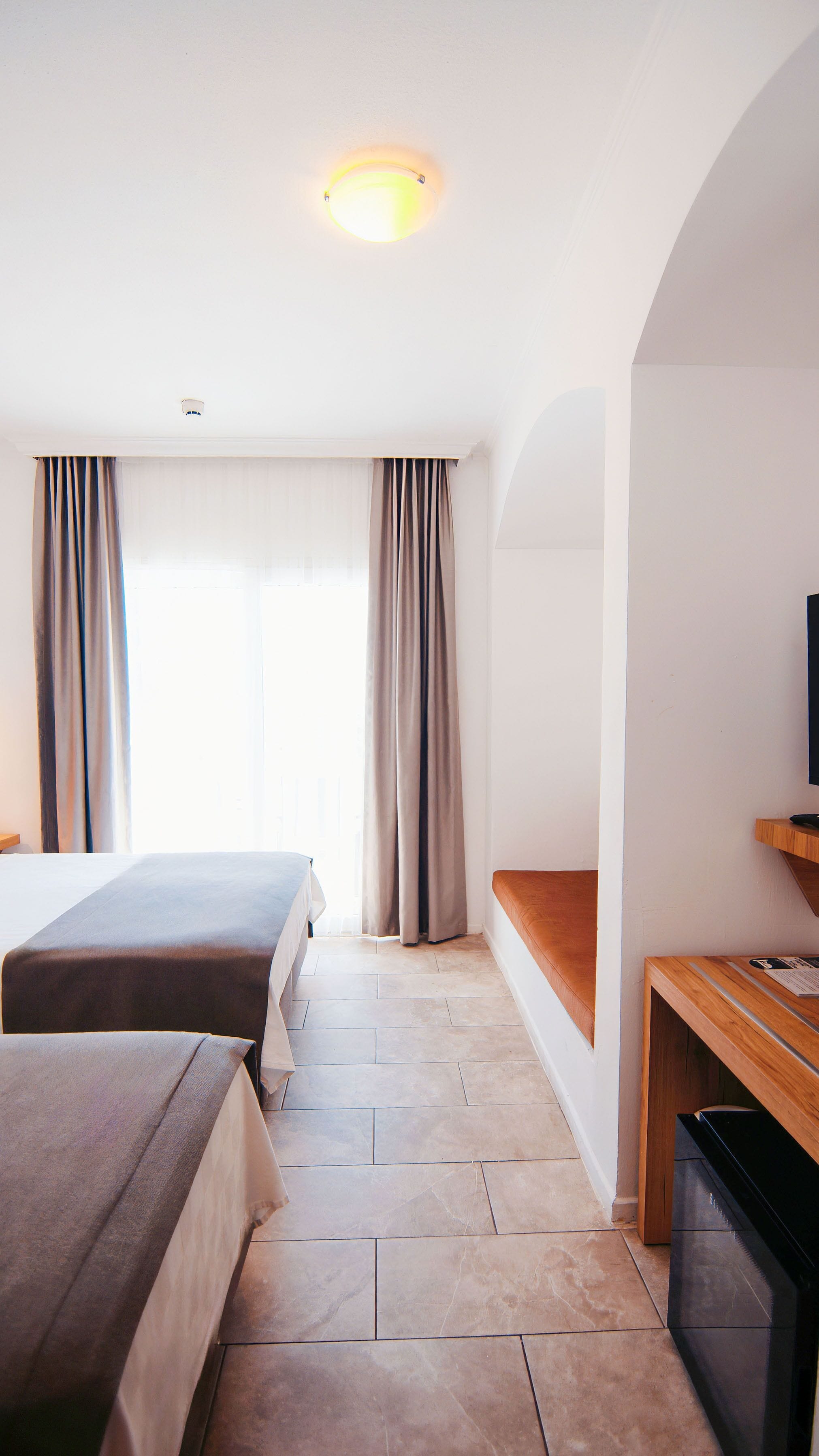 A hotel room with two beds, a television, a desk, and a large window with curtains.