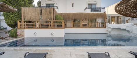 A pool area with lounge chairs, a thatched roof, and a building with a balcony.