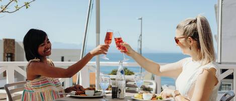 Two people toasting with drinks at an outdoor table with a view of the sea.