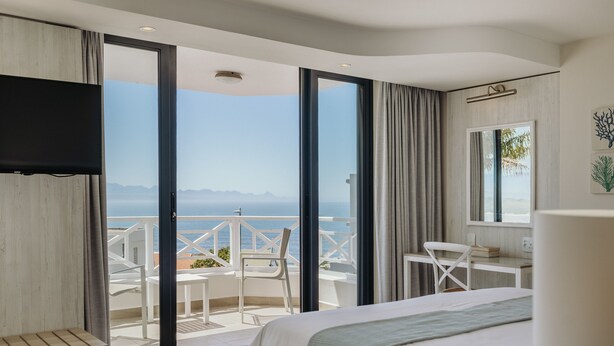 Une chambre d’hôtel avec un lit, un bureau, une chaise et une télévision. Elle dispose d’un balcon avec vue sur la mer et les montagnes.