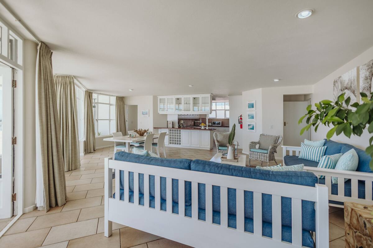 A spacious living area with a white sofa, a dining table, and a kitchen in the background.