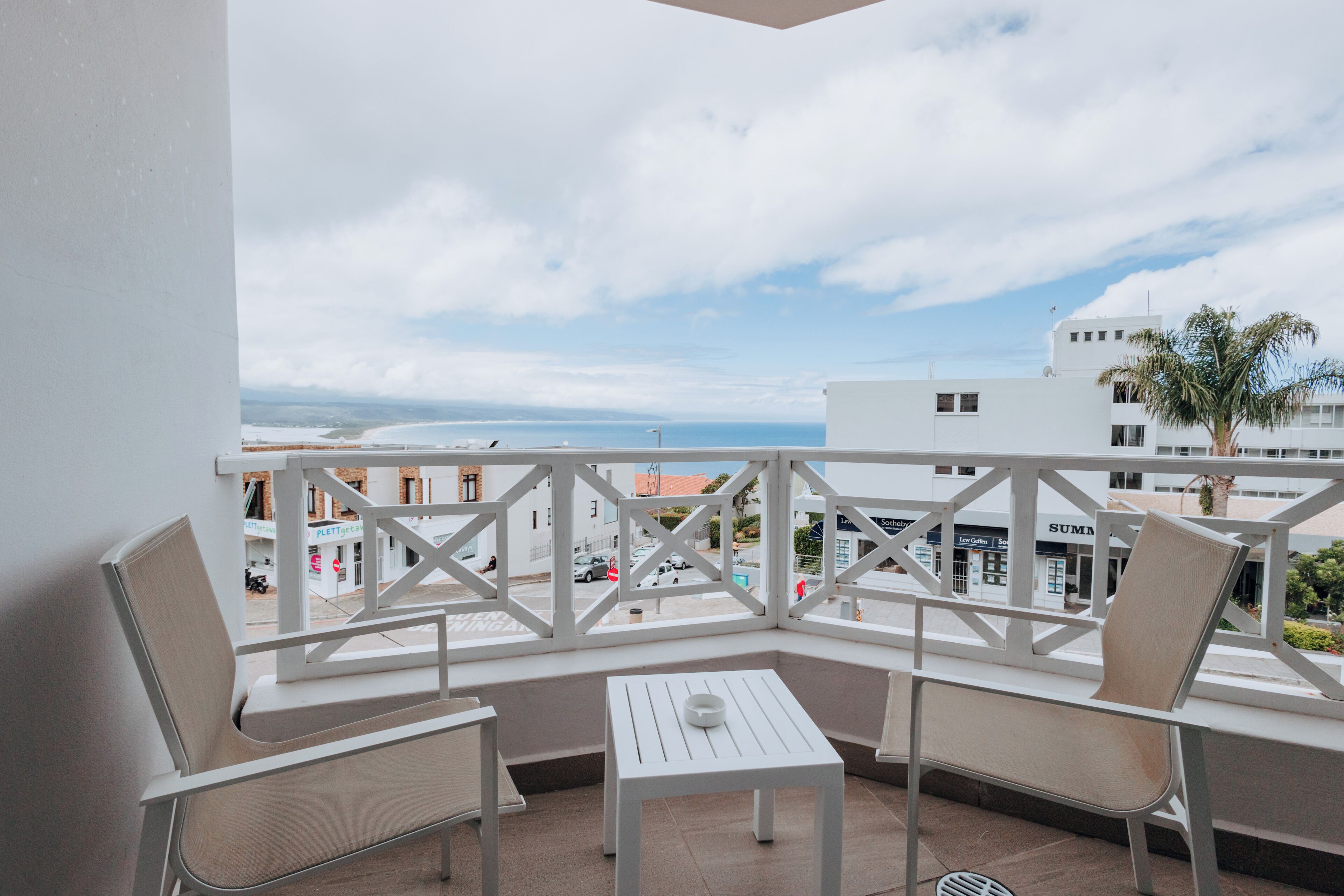 A balcony with two chairs and a small table, overlooking a coastal view with buildings and a palm tree.
