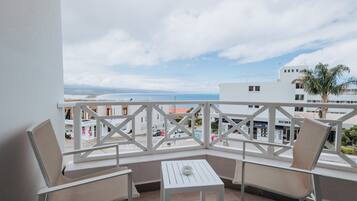 A balcony with two chairs and a small table, overlooking a coastal view with buildings and a palm tree.