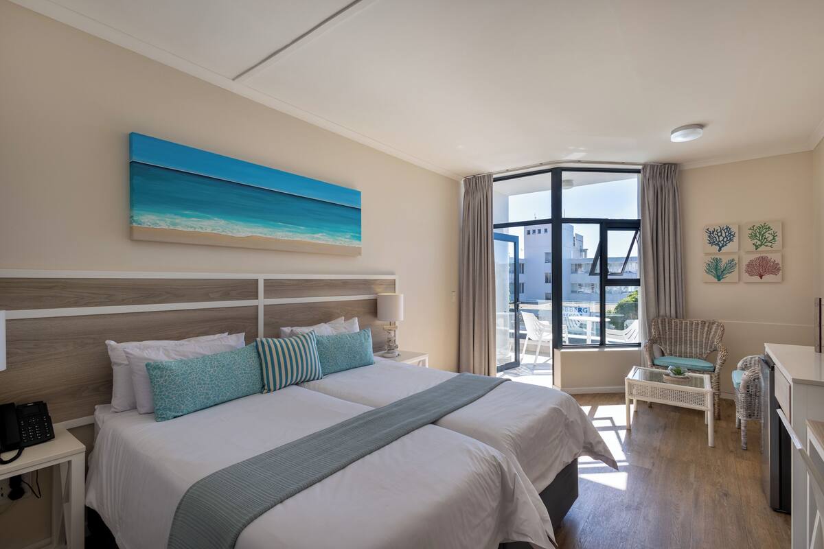 A hotel room with two beds, a large window, a view of the city, and a balcony with a chair.