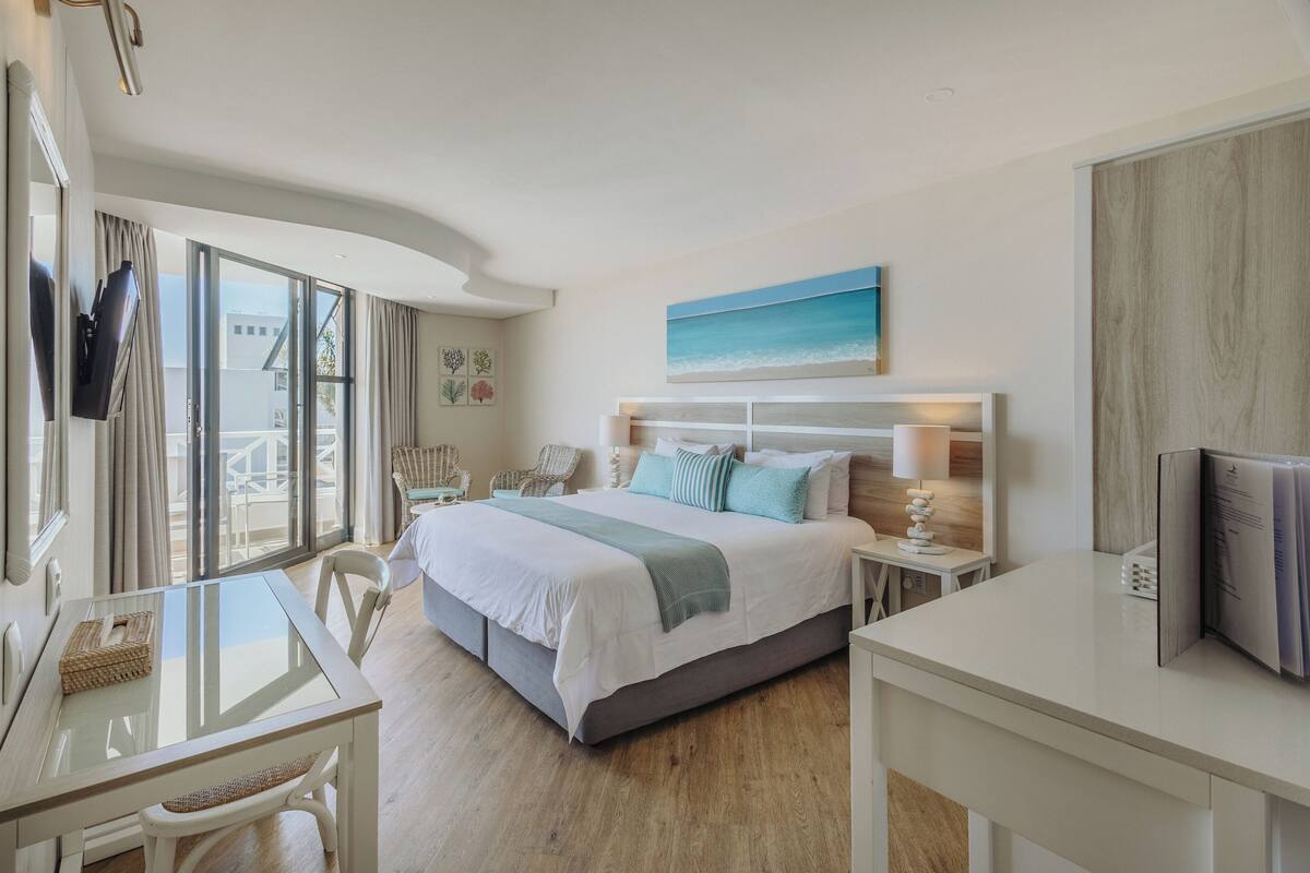 A hotel room with a large bed, a glass table, a chair, a desk, and a view of the outdoors.