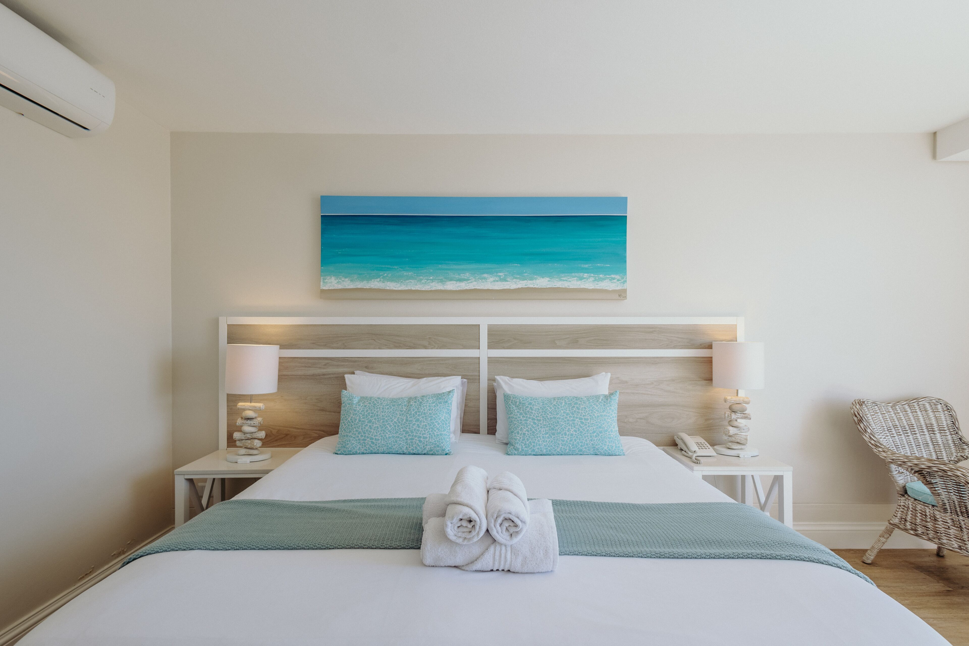 A neatly made bed with a view of a beach painting, a wicker chair, and a bedside lamp.