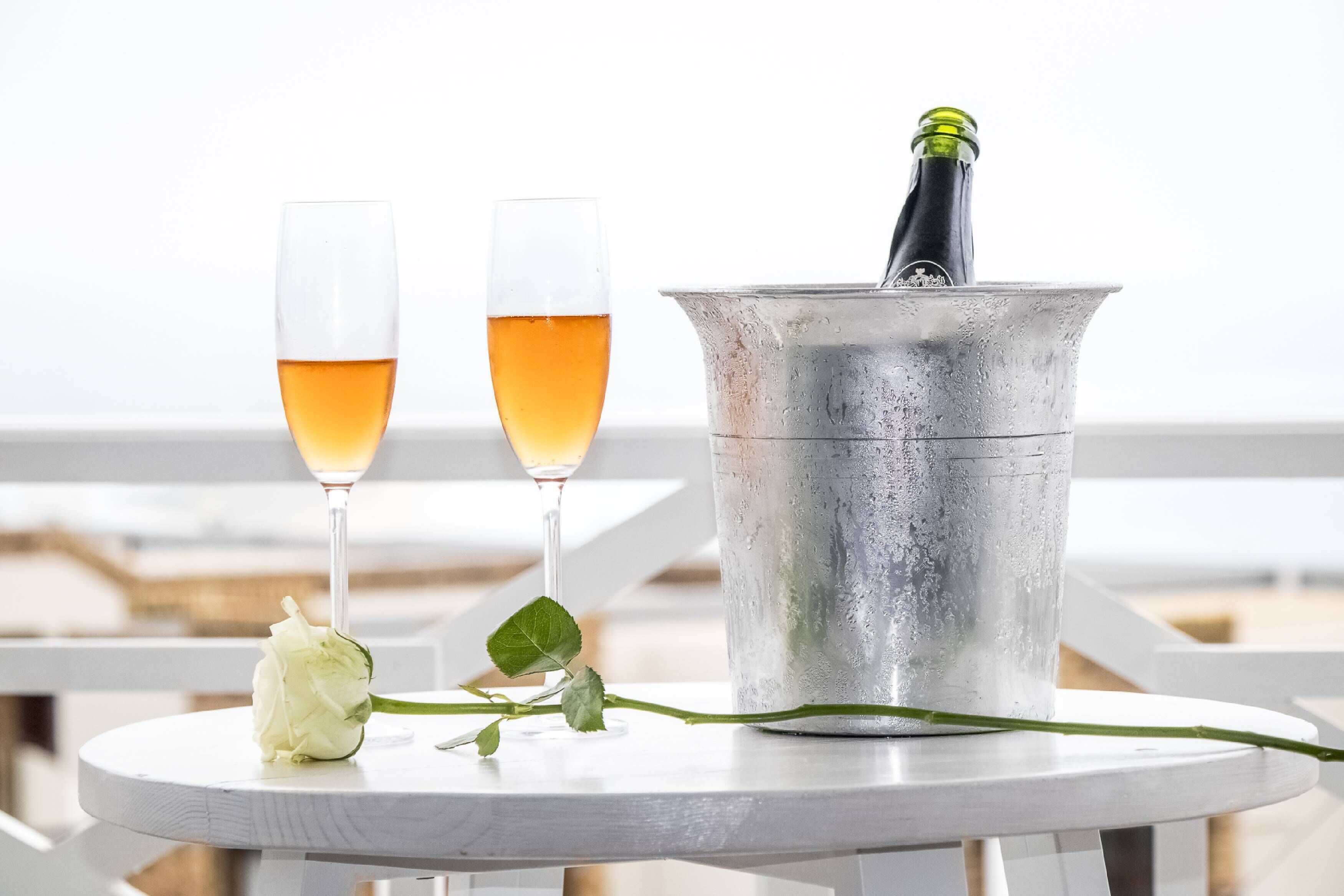 Dos copas de champán sobre una mesa con una botella en un cubo metálico, una rosa blanca y vista al mar.