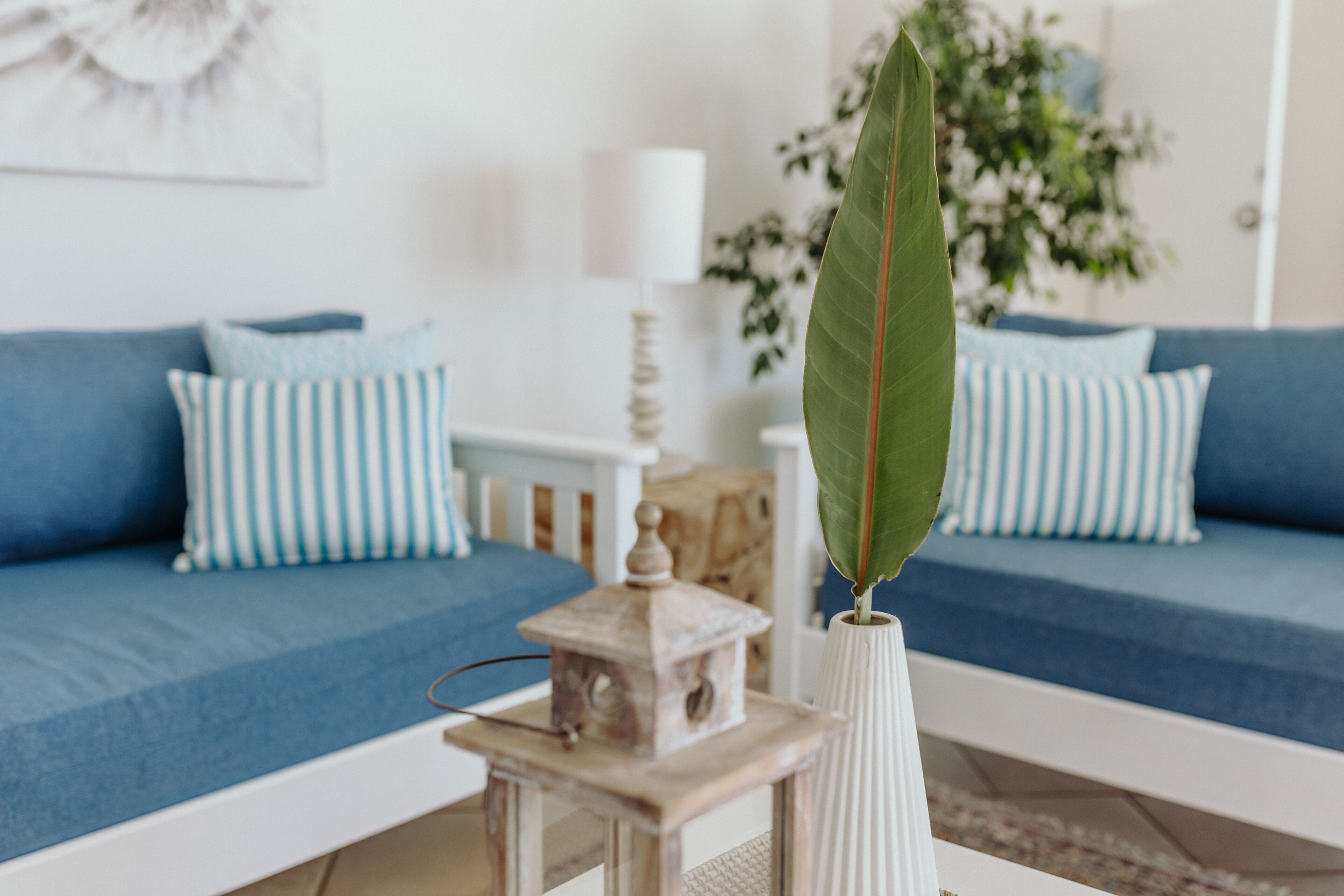 A living room with a blue sofa, striped pillows, a white vase with a green leaf, and a small wooden lantern.