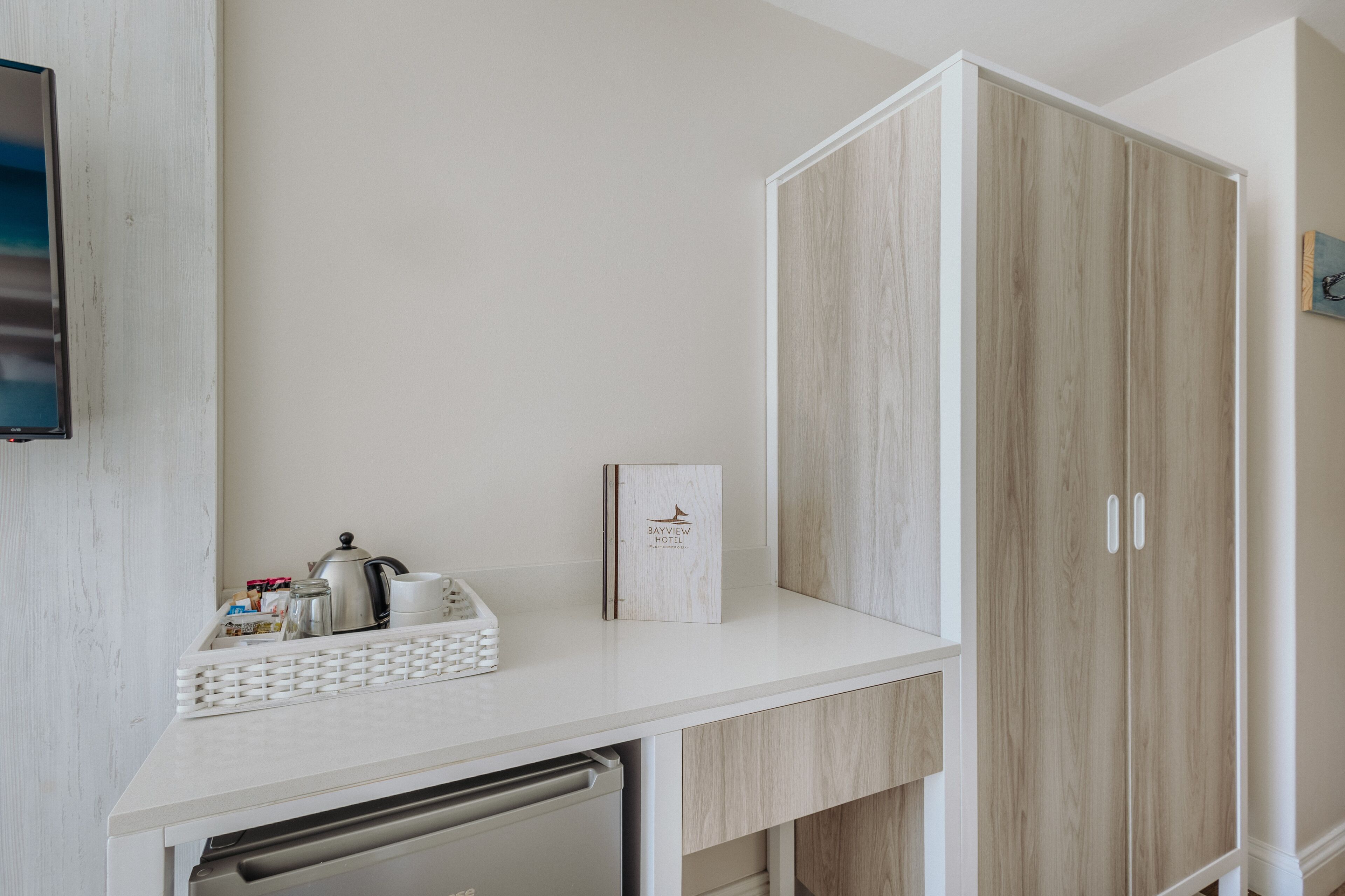 Une chambre d’hôtel avec un comptoir blanc, une armoire en bois, une télévision et une petite table sur laquelle se trouvent un panier et une bouilloire.