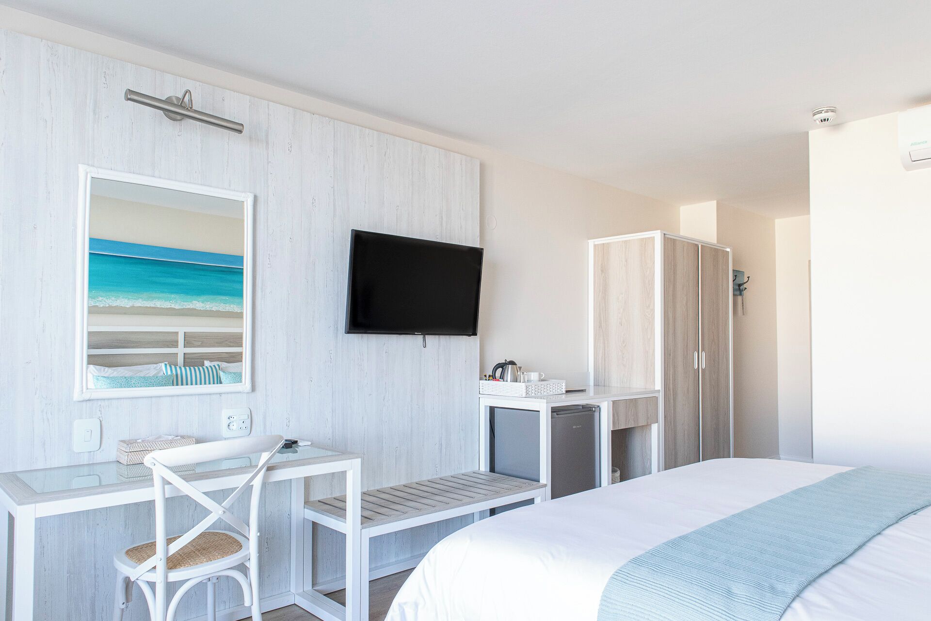 A hotel room with a bed, a desk with a chair, a television, a wardrobe, and a small kitchenette.