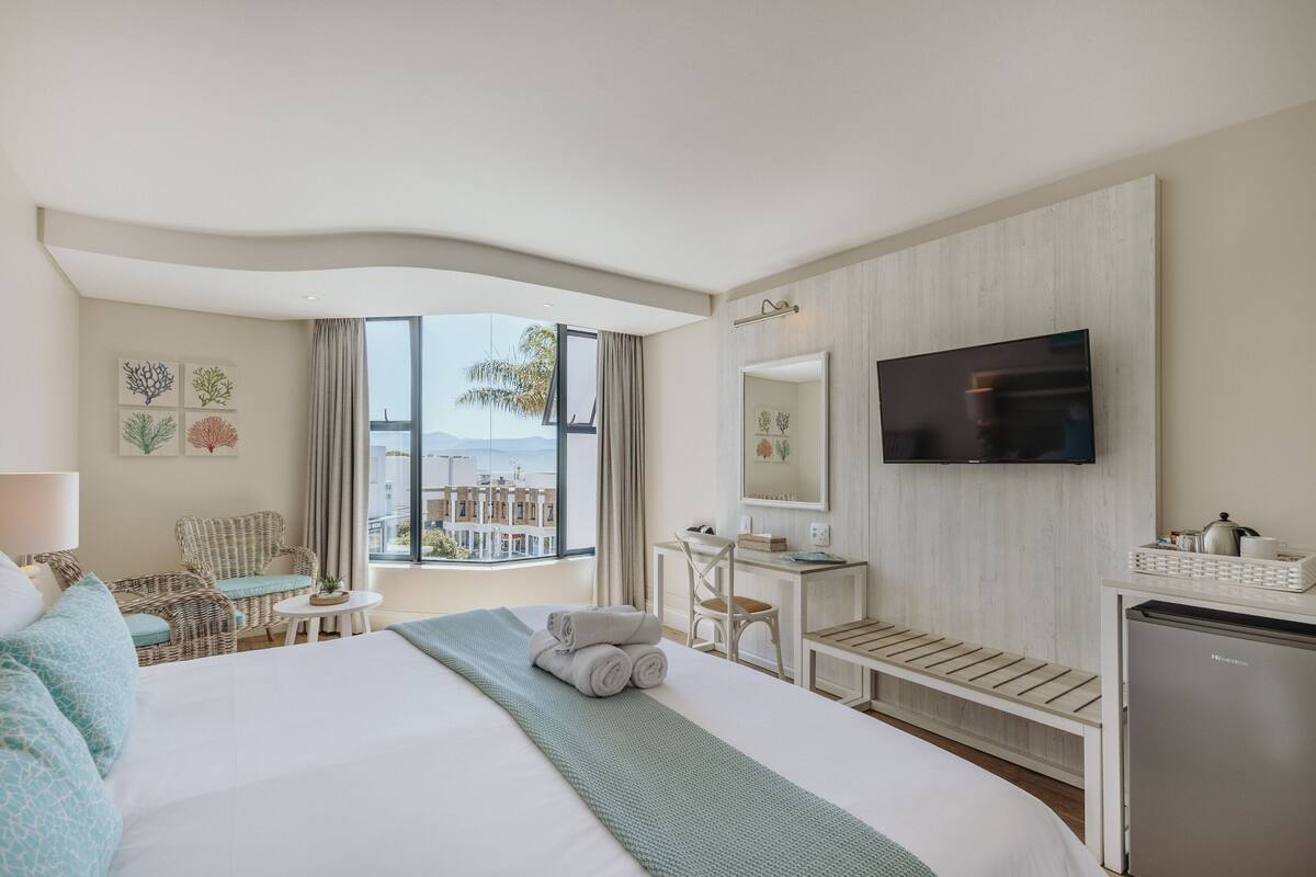 A hotel room with a large bed, a desk, a chair, a TV, and a view of buildings through the window.
