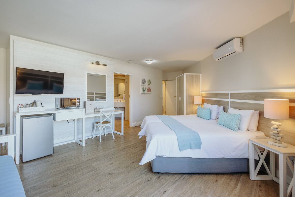 A modern hotel room with a bed, desk, and chair, a flat-screen TV, and an air conditioning unit.