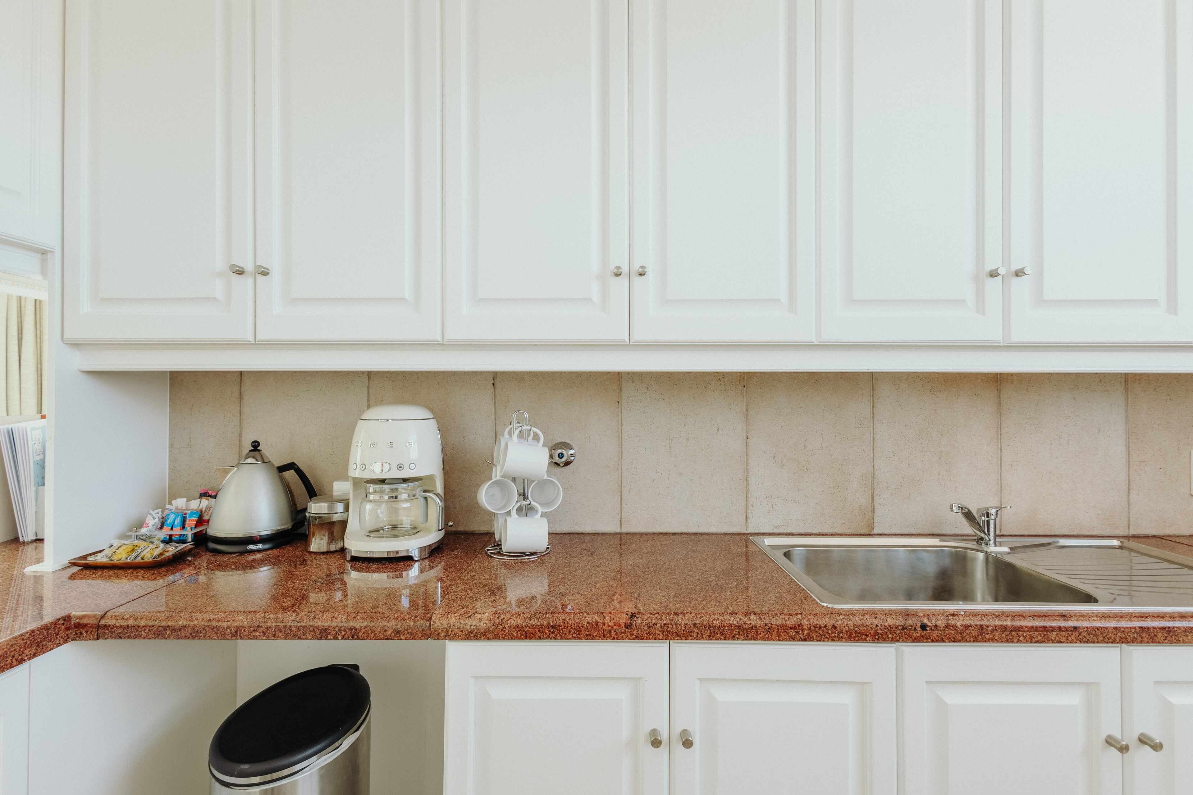 A kitchen with white cabinets, a granite countertop, a coffee maker, a kettle, and a sink.