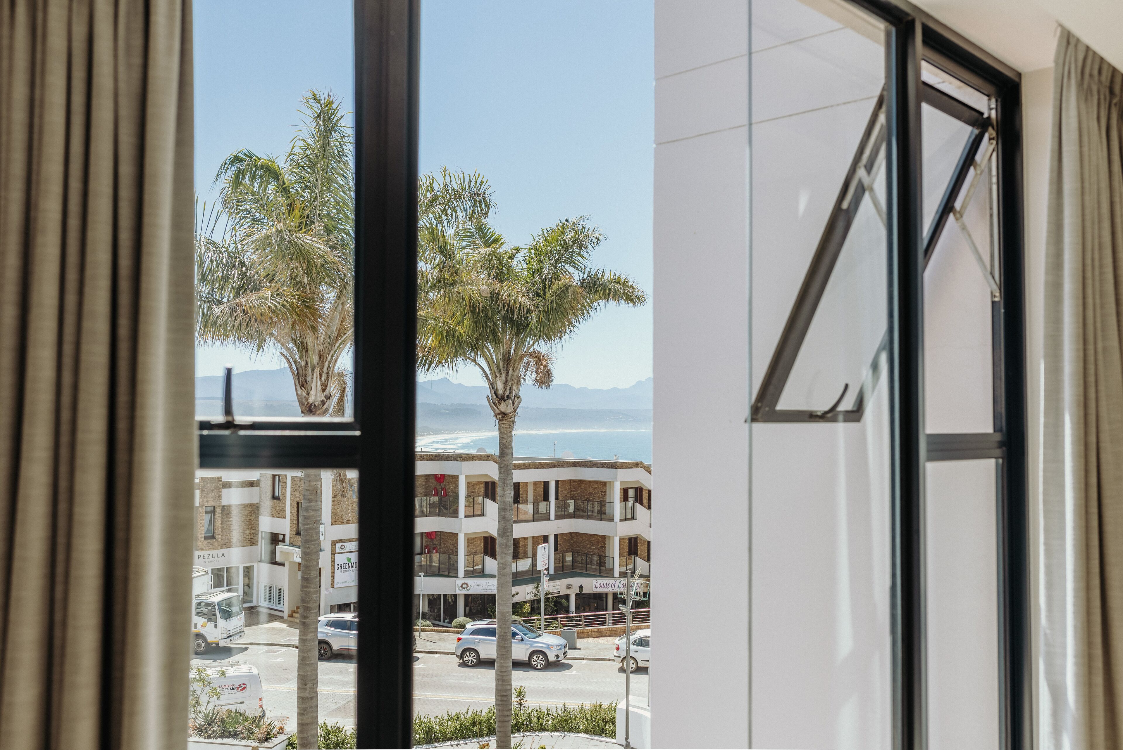 A view from a window overlooking a street with palm trees, a building with multiple floors, and a clear sky.