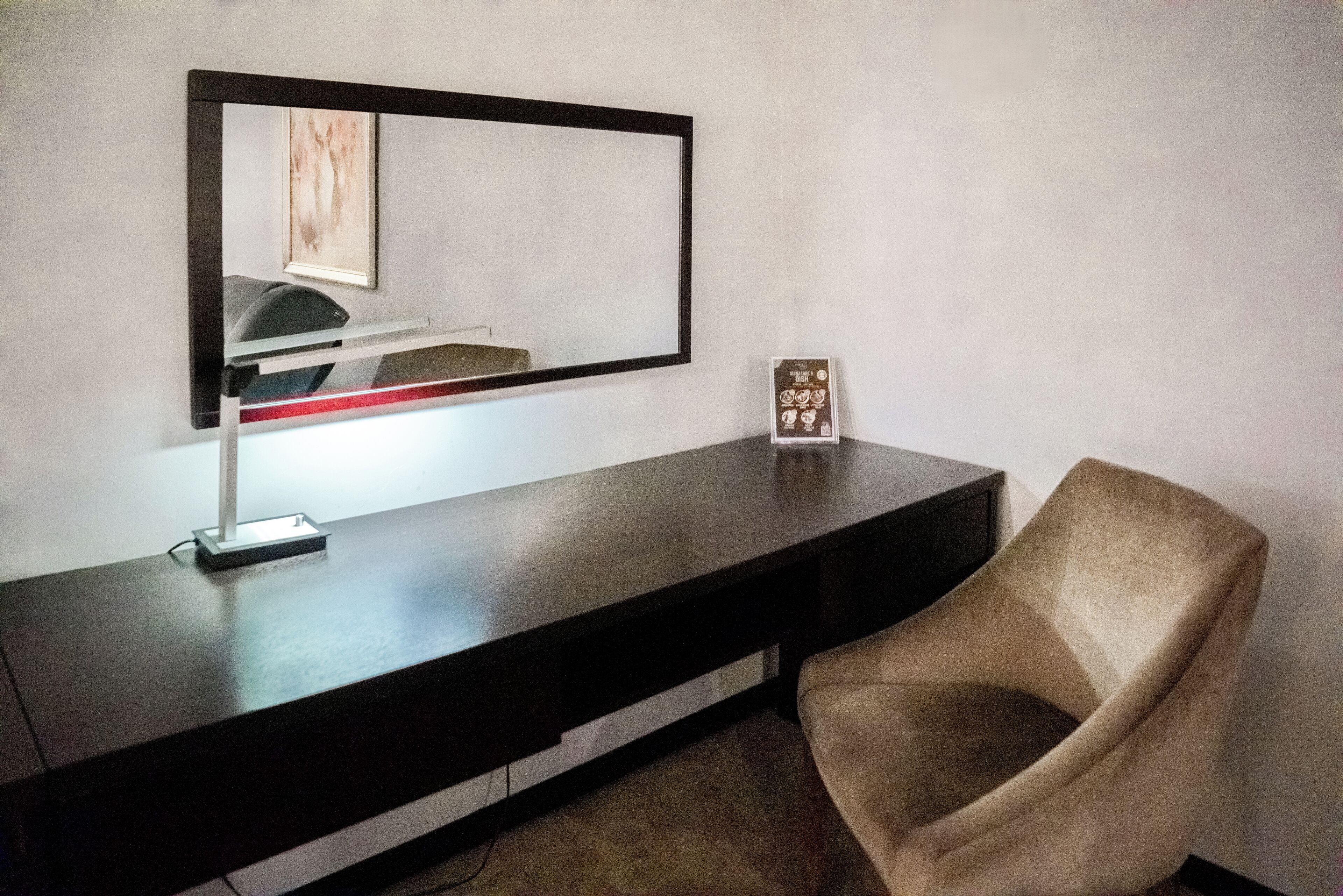 A modern room with a dark wooden desk, a chair, a large mirror, and a framed picture on the wall.