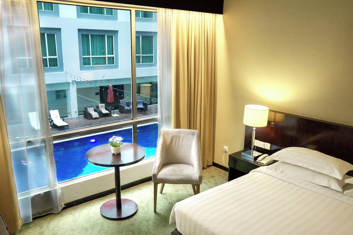 Habitación de hotel con cama, una silla, una mesita con una planta, vistas a la piscina y una lámpara.