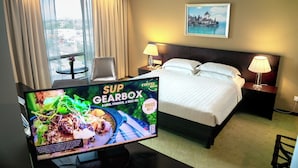 Ein Hotelzimmer mit einem Bett, zwei Nachttischen mit Lampen, einem Fernseher, der eine Speisekarte zeigt, einem Fenster mit Vorhängen und einem Gemälde an der Wand.