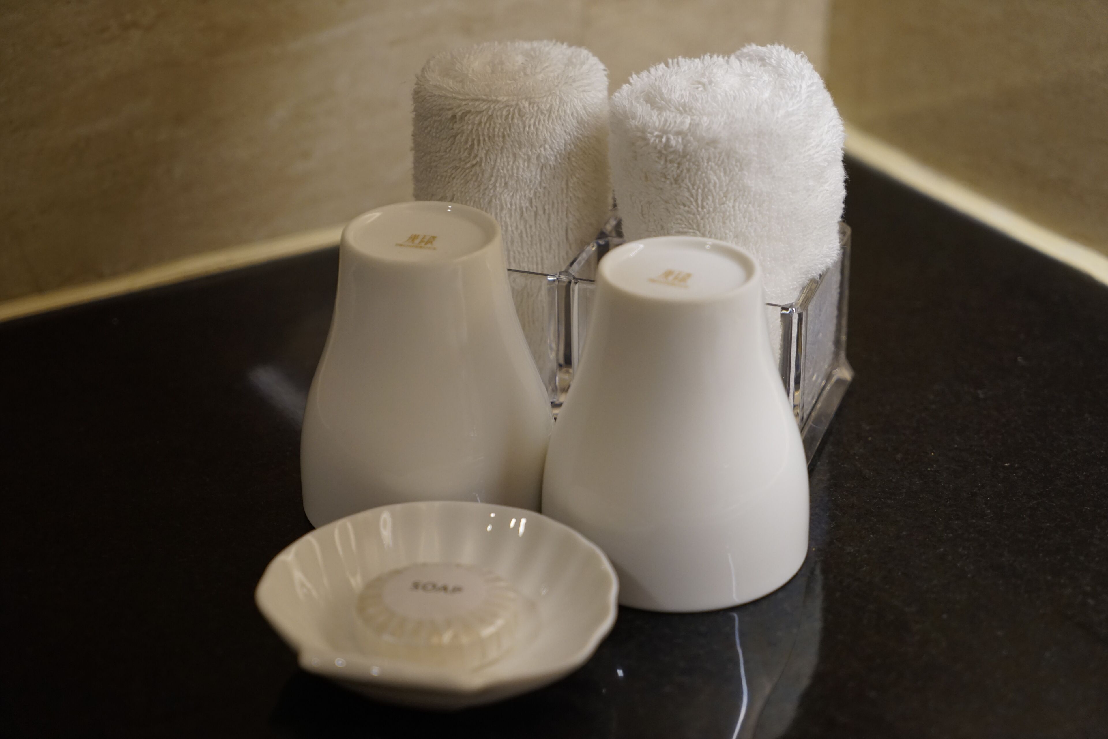 A bathroom with a black countertop, three white bottles with soap labels, and two rolled towels in a holder.