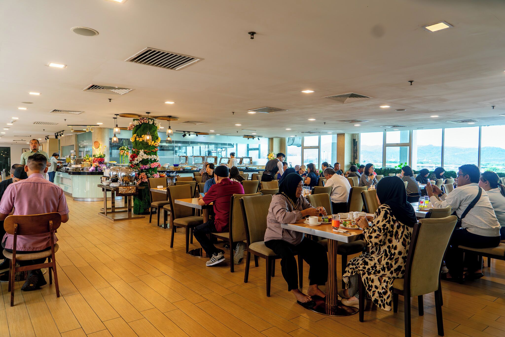 A spacious dining area with multiple tables and chairs, large windows offering a view of the outdoors, and a central decorative floral arrangement.