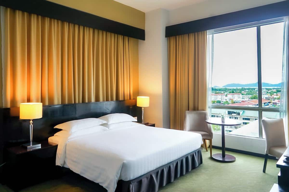 Habitación de hotel con una cama grande, dos lámparas de noche, una silla, una mesita y vistas a un paisaje urbano a través de amplias ventanas.