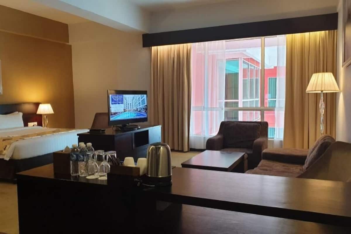 Habitación de hotel con una cama, un escritorio con computadora, televisor, cafetera y vista a un edificio a través del ventanal.
