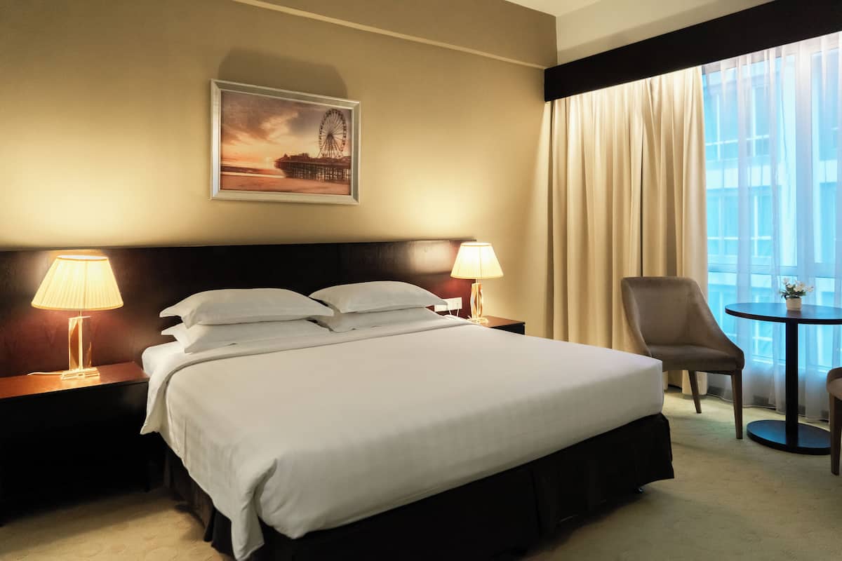 Habitación de hotel con una cama grande, dos lámparas de noche, una silla, una mesita y una ventana con cortinas.