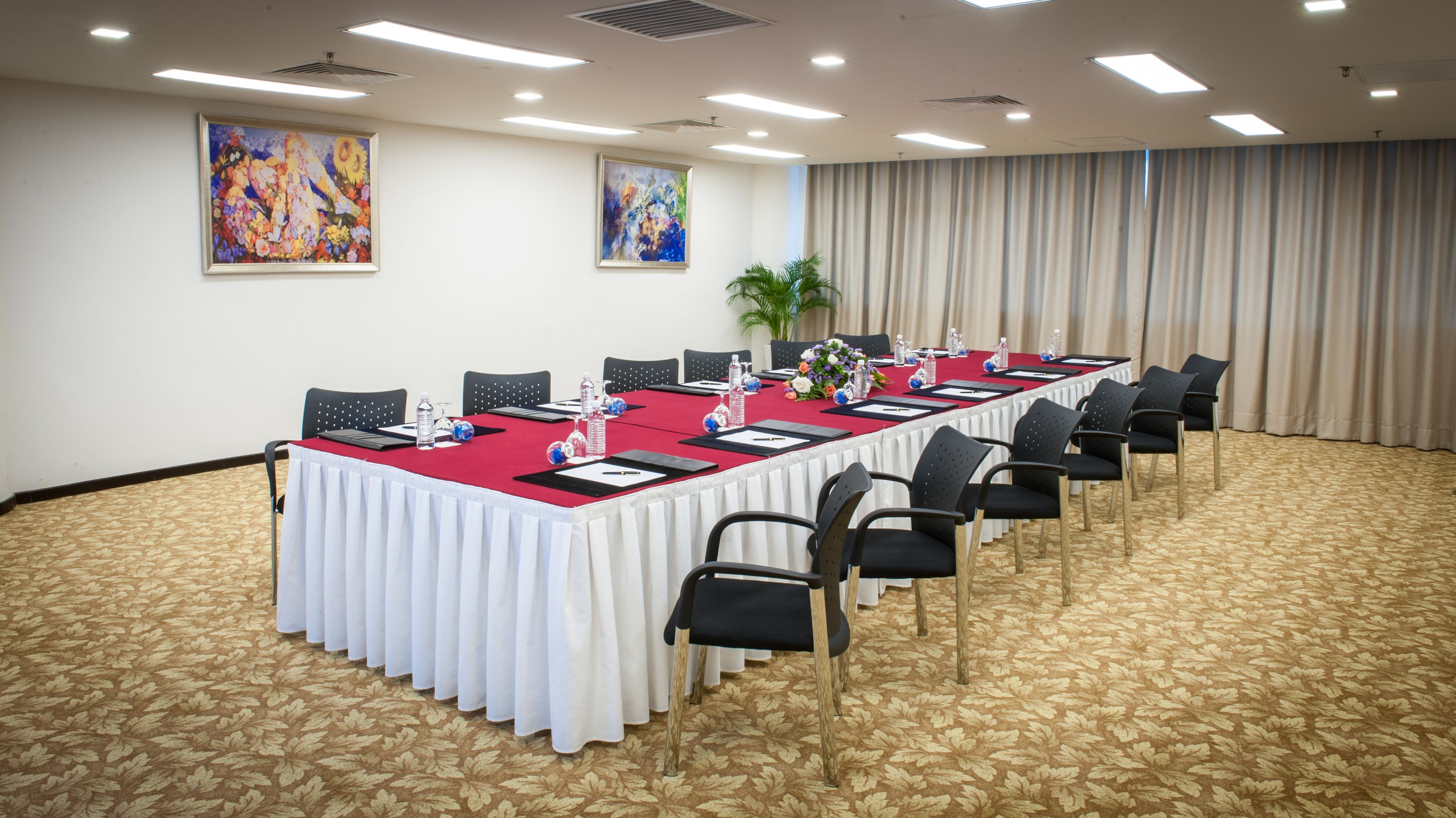 A conference room with a long table, black chairs, and a red tablecloth. There are two paintings on the wall and a plant in the corner.