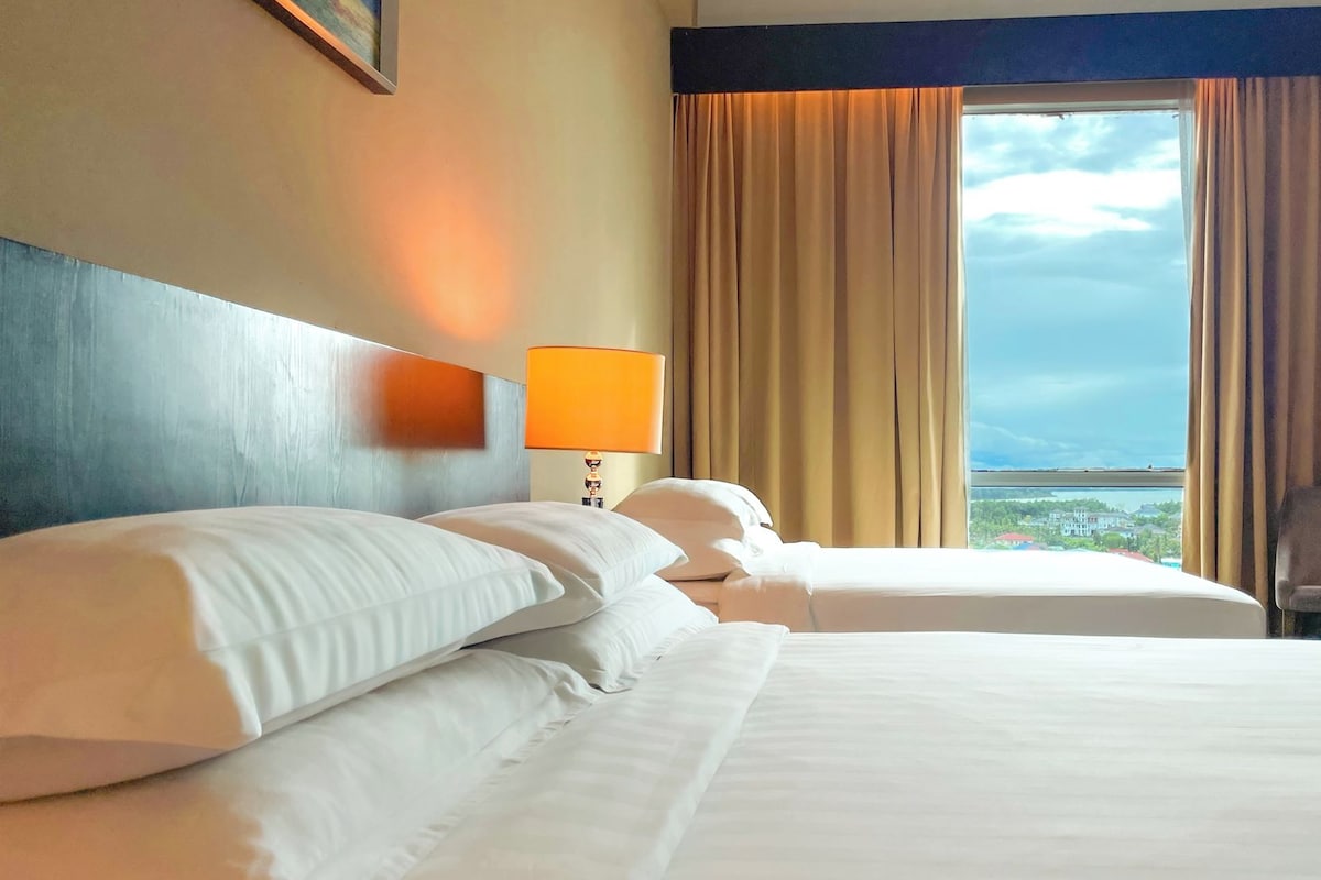 Habitación de hotel con dos camas, una mesita de noche con lámpara y vistas a la ciudad a través de amplios ventanales.