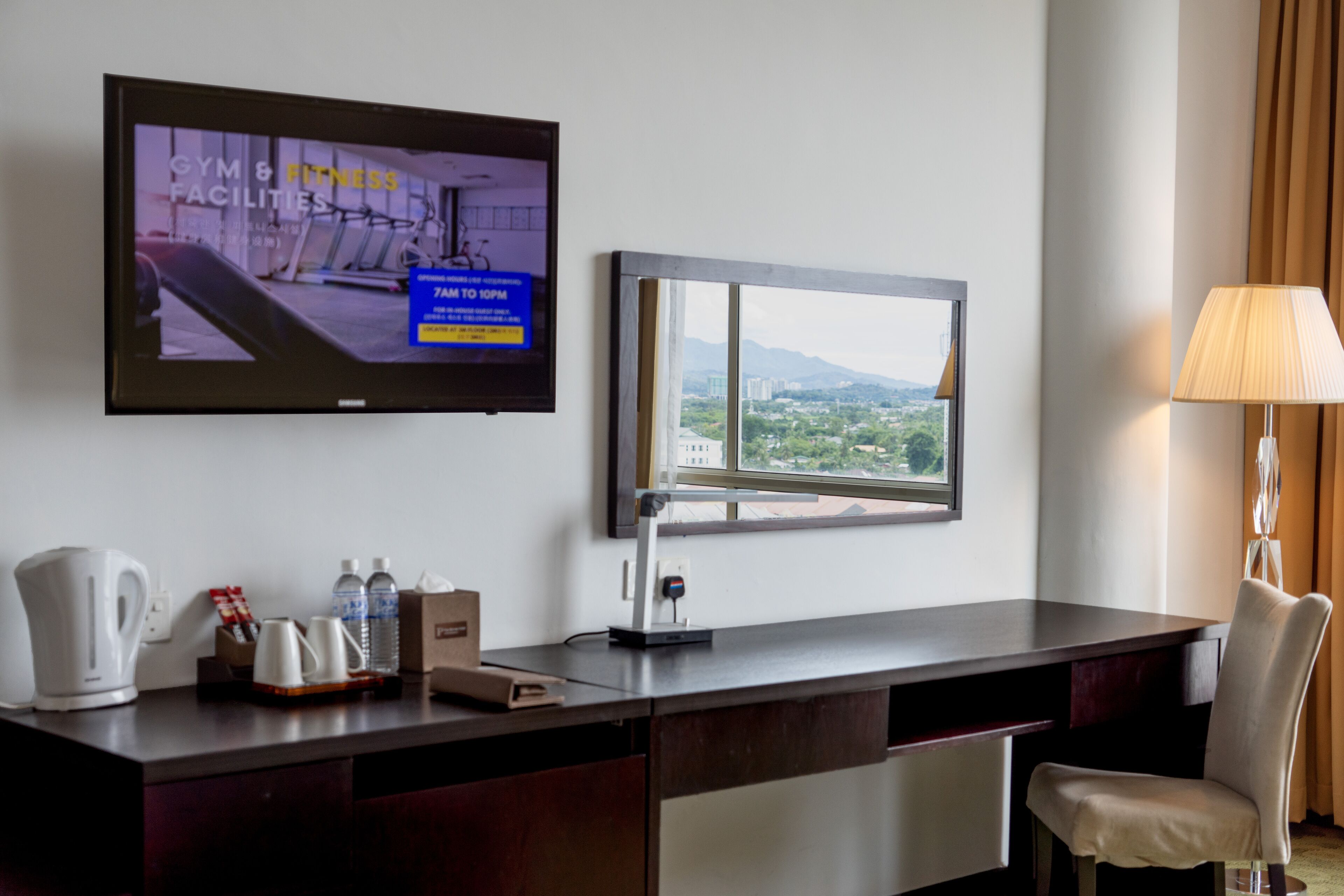 A hotel room with a desk, a television displaying gym and fitness facilities, a window with a city view, and a lamp.