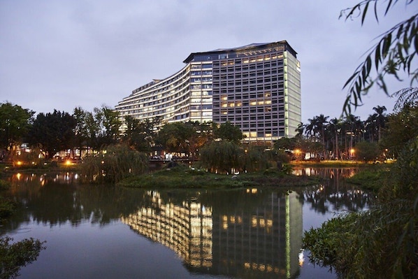 Un hôtel moderne à la façade incurvée, qui se reflète dans une étendue d’eau paisible, et qui est entouré de verdure et de palmiers.