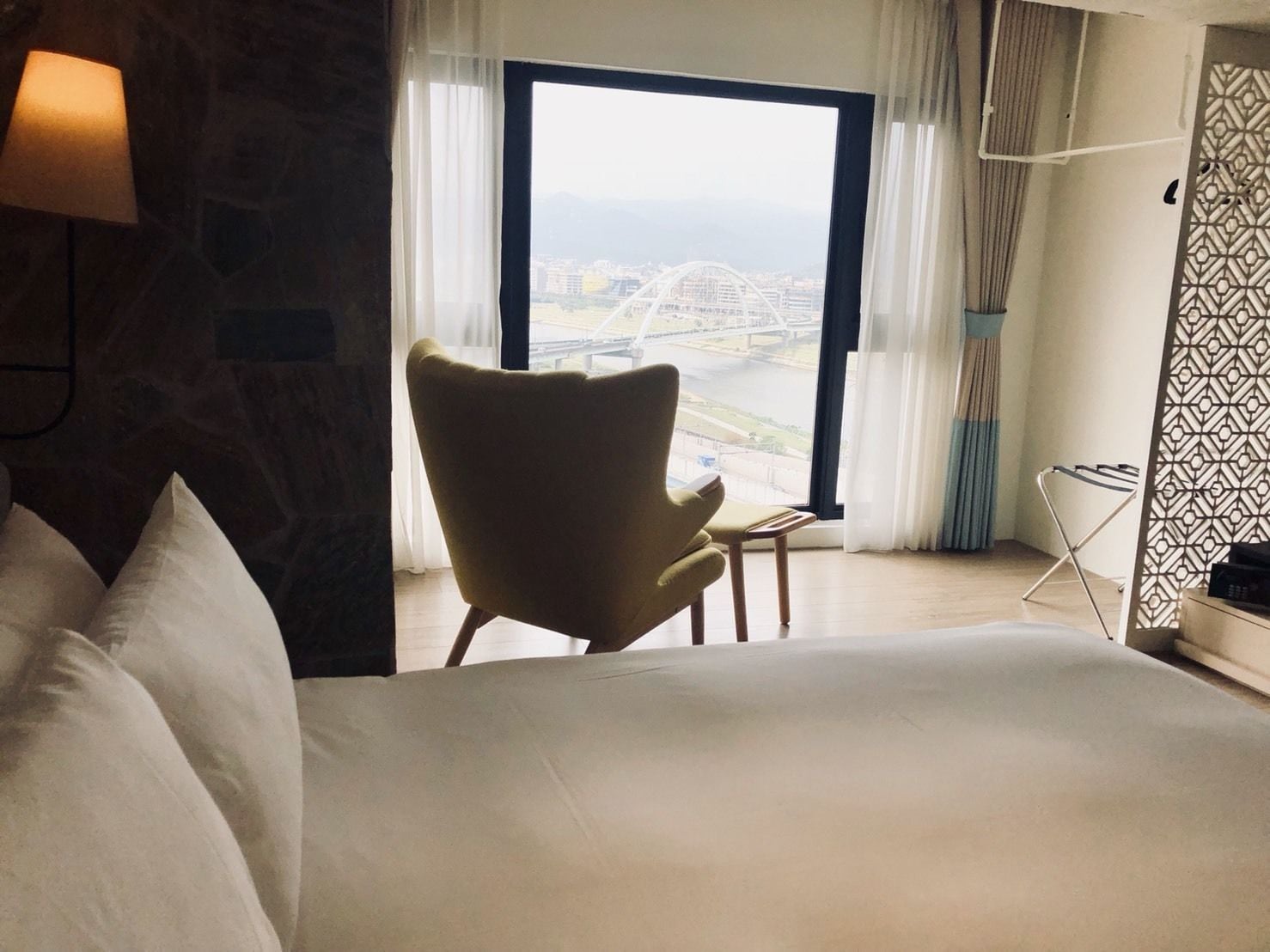 A hotel room with a large bed, a chair, a view of a bridge, and a window with curtains.